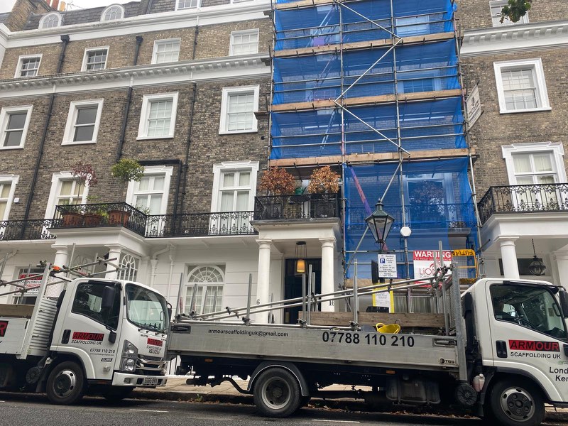 Terraced stucco house wrapped in blue sheeted scaffold with an Armour lorry showing the phone number